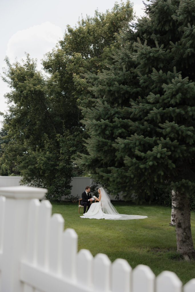 Bride and groom | sitting together reading vows before their ceremony | backyard wedding with white fence